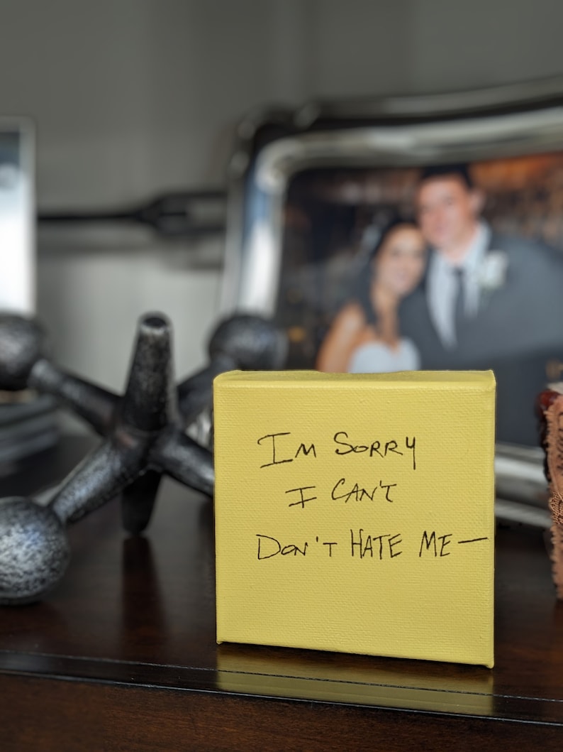 May include: A yellow sticky note with the handwritten message "I'm sorry I can't Don't hate me" on a wooden surface. A blurry photo of a couple is in the background.
