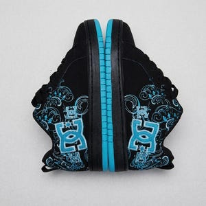 Op de afbeelding: Een paar zwarte sneakers met een felblauw DC-logo en ingewikkeld blauw krulwerk-borduurwerk op de zijkanten. De schoenen hebben zwarte veters, zwarte zolen en een opvallende blauwe streep langs de rand van de rubberen buitenzolen.