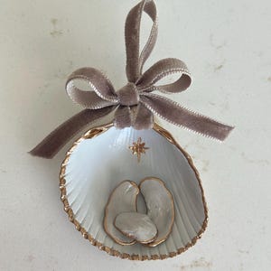 May include: A white seashell ornament with a gold-painted rim, featuring a small gold star icon and three white clay figures inside. The shell is topped with a taupe velvet ribbon tied in a bow, creating a decorative hanging piece.