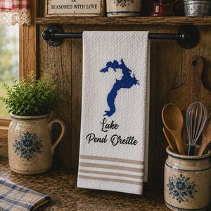 May include: A white waffle-weave kitchen towel hanging on a black metal rod. The towel features a blue graphic of Lake Pend Oreille, the text "Lake Pend Oreille" in black script, and three horizontal tan stripes near the bottom edge of the fabric.