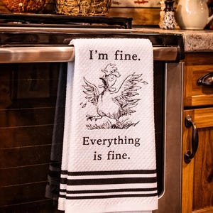 May include: White kitchen towel with black stripes and the text "I'm fine. Everything is fine." A cartoon chicken illustration is featured on the towel. The towel is hanging on an oven door.