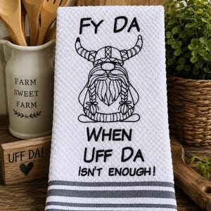 May include: A white waffle-weave kitchen towel featuring black embroidery of a Viking gnome with braided hair and horns. The text reads FY DA WHEN UFF DA ISN'T ENOUGH! The bottom of the towel has three horizontal gray stripes.