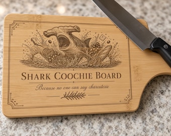 May include: A rectangular bamboo cutting board with a laser-engraved illustration of a hammerhead shark emerging from water with food items. The text reads SHARK COOCHIE BOARD and Because no one can say charcuterie. A kitchen knife rests on the board.