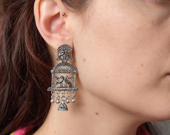 May include: A silver-toned dangle earring featuring an oxidised finish with intricate metalwork. The design includes a top arch, a central rectangular frame with two bird figures, and a bottom row of small hanging beads and a bell charm.