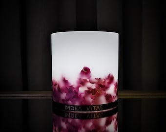 Può includere: Un portacandele cilindrico bianco illuminato dall'interno, caratterizzato da una base decorata con petali di fiori rosa scuro e viola incastonati. Una fascia nera sul fondo riporta la scritta bianca MORA VITAE in maiuscolo.