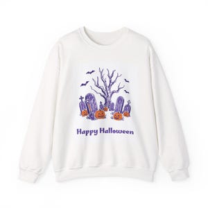 Puede incluir: Una sudadera blanca de manga larga con un estampado gráfico en morado y naranja de un cementerio con un árbol desnudo, murciélagos, calabazas y una pala. El texto "Happy Halloween" aparece impreso en color morado debajo.