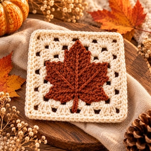 May include: A square crochet granny square featuring a cream-colored border with a textured, dark brown maple leaf motif appliqued in the center. The piece is crafted with yarn and displays a detailed, raised leaf pattern on a neutral background.