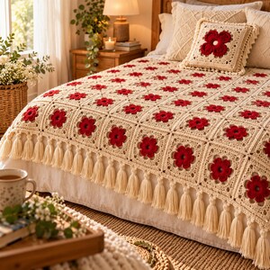 May include: A cream-colored crochet blanket featuring a repeating pattern of dark red flower motifs in each square, finished with a decorative fringe of long cream tassels along the edge, paired with a matching square throw pillow.