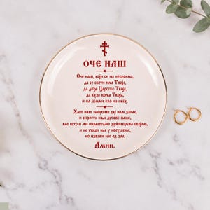 May include: A round white ceramic plate with a gold-toned rim, featuring a red Orthodox cross and the Lord's Prayer in Serbian Cyrillic text printed in red. The plate is displayed alongside a pair of small gold hoop earrings.