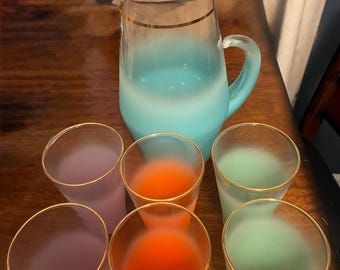 May include: A glass pitcher with a light blue frosted base and a gold rim, accompanied by six matching frosted glasses in purple, orange, and green, each featuring a gold rim. The set includes two glasses of each color, all with a smooth finish.