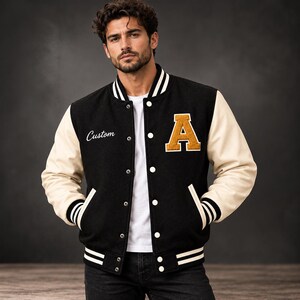 May include: A black varsity jacket with cream-coloured sleeves, featuring a large gold letter A patch on the left chest and the word Custom embroidered in white script on the right chest. The jacket has a snap-button front and striped trim.