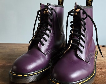 Peut inclure: Une paire de bottines en cuir violet avec des lacets noirs, huit œillets et des coutures jaunes le long de la semelle. Chaque botte présente une languette noire au talon avec le texte jaune AirWair, un bout rond et une semelle en caoutchouc noir.