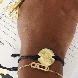 May include: A gold bracelet with a black cord and a round charm that says "Find the magic". The charm is attached to the cord with a gold chain. The bracelet also has a smaller gold chain with a rectangular charm that has three small white stones.