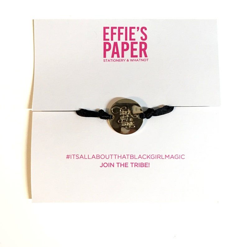 May include: A black string bracelet with a gold charm that says "Black Girl Magic". The bracelet is on a white card with the text "EFFIE'S PAPER STATIONERY & WHATNOT #ITSALLABOUTTHATBLACKGIRLMAGIC JOIN THE TRIBE!"