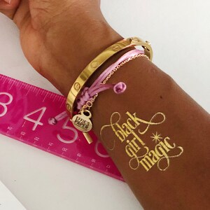 May include: A gold bracelet with a pink string bracelet and a charm that says "Effie's Paper" on a person's arm with a gold temporary tattoo that says "Black Girl Magic".