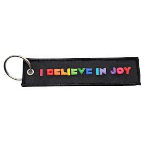 Può includere: Portachiavi rettangolare nero con anello argentato. Il portachiavi presenta la scritta ricamata "I BELIEVE IN JOY" nei colori dell'arcobaleno. Un accessorio semplice e colorato.