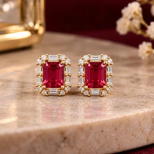 May include: A pair of gold stud earrings featuring a central emerald-cut red gemstone surrounded by a halo of alternating baguette and round white stones. The earrings are set in a yellow gold metal frame with a polished finish.