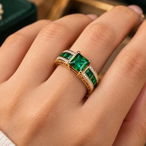 May include: A gold ring featuring a large square-cut green gemstone in the center, flanked by smaller square green stones on each side. The band is accented with rows of tiny white stones and intricate filigree metalwork detailing.