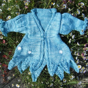 Op de afbeelding: Een lichtblauwe gebreide cardigan met een gegolfde rand en witte bloemenversieringen. De cardigan is open aan de voorkant en heeft een V-hals.