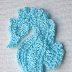 May include: A crocheted seahorse made of light blue yarn. The seahorse has a detailed head and a curled tail.