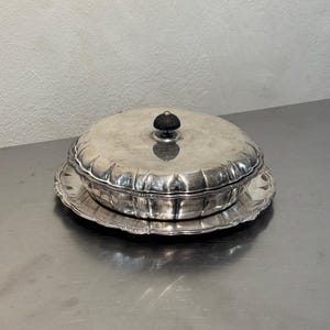 Puede incluir: Un plato de servicio redondo de metal plateado con tapa a juego y borde festoneado. La tapa presenta un pequeño pomo oscuro y redondeado en el centro. El plato descansa sobre una bandeja decorativa ancha de metal plateado.