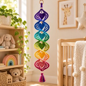 May include: A hanging spiral mobile featuring a vertical sequence of six crocheted hearts in rainbow colors, including purple, blue, teal, green, yellow, and pink. The structure is accented with small wooden beads and a multi-colored tassel.