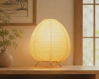 May include: A table lamp with a rounded, off-white paper shade featuring horizontal ribbing. The lamp is supported by three thin, gold-toned metal legs that elevate the shade above the surface, emitting a warm, soft glow.