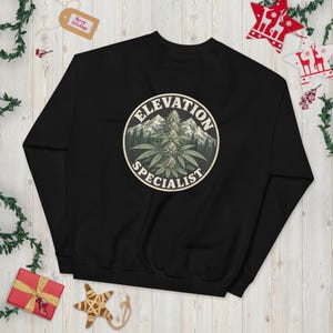 Könnte beinhalten: Ein schwarzes Langarm-Sweatshirt mit einer kreisförmigen Grafik auf der Brust. Die Grafik zeigt eine Cannabispflanze vor einer Bergkette und einem Wald, mit dem Text "ELEVATION SPECIALIST" in einem Bogen um den kreisförmigen Rand.