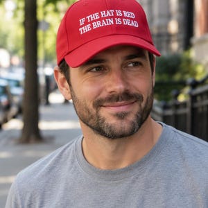 May include: A bright red baseball cap featuring white embroidered text on the front that reads, IF THE HAT IS RED THE BRAIN IS DEAD. The cap has a structured crown, a curved brim, and a button on top, constructed from a solid fabric material.