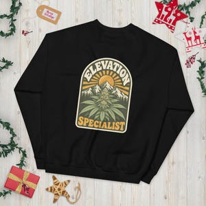 Könnte beinhalten: Ein schwarzes Langarm-Sweatshirt mit einer zentralen Grafik einer Cannabispflanze vor Berggipfeln und einer Sonne. Der Text "ELEVATION SPECIALIST" ist in einer fetten, gelben Retro-Schriftart bogenförmig um die Illustration angeordnet.