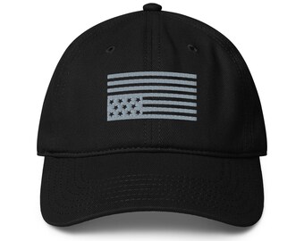 May include: A black baseball cap featuring an embroidered light gray graphic of the United States flag on the front panel. The design consists of horizontal stripes and a rectangular field containing stars, all rendered in a monochromatic gray tone.