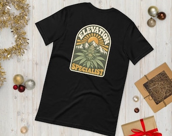 May include: A black short-sleeve t-shirt featuring a graphic print on the back. The design shows a mountain range under a sun, with a cannabis plant in the foreground and the text "ELEVATION SPECIALIST" arched within a frame.