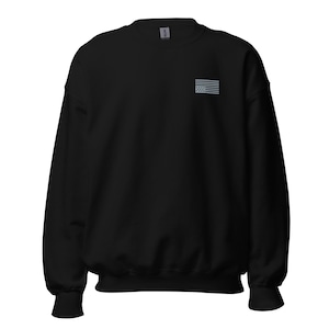 Könnte beinhalten: Ein schwarzes Langarm-Sweatshirt mit Rundhalsausschnitt und einer kleinen, gestickten grauen amerikanischen Flagge auf der linken Brustseite. Das Kleidungsstück verfügt über einen klassischen gerippten Kragen, Bündchen und Saum sowie ein sichtbares Markenetikett am Hals.