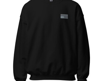May include: A black long-sleeve crewneck sweatshirt featuring a small, embroidered grey American flag graphic positioned on the upper left chest area. The garment has a standard ribbed collar, cuffs, and hem, with a visible brand label at the neck.