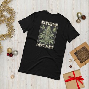 Könnte beinhalten: Ein schwarzes Kurzarm-T-Shirt mit einer rechteckigen Grafik auf der Rückseite. Die Grafik zeigt eine botanische Illustration einer Cannabispflanze mit dem Schriftzug "ELEVATION SPECIALIST" in weißer Serifenschrift.