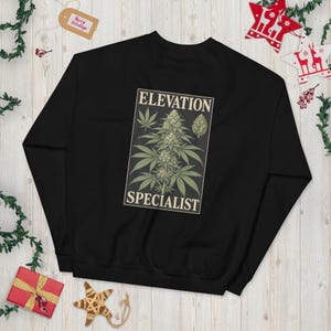 Könnte beinhalten: Ein schwarzes Langarm-Sweatshirt mit einer rechteckigen Grafik auf der Brust. Die Abbildung zeigt eine grüne Cannabispflanze mit dem Text "ELEVATION SPECIALIST" in einer cremefarbenen Serifenschrift oberhalb und unterhalb des Motivs.
