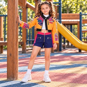 May include: A cropped orange jacket with black trim, spiked shoulders, and colorful cloud and wave patterns, paired with a white mock-neck top and navy blue shorts featuring a front zipper, pink side panels, and white sneakers.