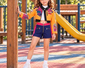 May include: A cropped orange jacket with black trim, spiked shoulders, and colourful cloud and wave patterns, paired with a white mock-neck top and navy blue shorts featuring a front zip, pink side panels, and white trainers.
