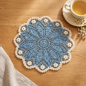 Könnte beinhalten: Ein rundes, filigranes Häkeldeckchen mit einem zentralen blauen Blumenmuster und strahlenförmigen Blütenblättern, eingefasst von einem weißen, gewellten Rand, der mit kleinen, gleichmäßig verteilten weißen Häkelblüten verziert ist.