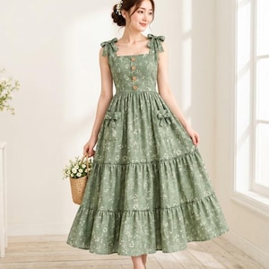 Könnte beinhalten: Ein salbeigrünes Midikleid mit weißem Blumenmuster, eckigem Ausschnitt und zu Schleifen gebundenen Schulterträgern. Das Oberteil hat eine vertikale Reihe aus vier braunen Knöpfen und der Rock verfügt über zwei aufgesetzte Taschen mit kleinen Schleifen.