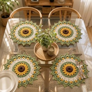 Könnte beinhalten: Vier runde Häkel-Platzsets mit Sonnenblumenmuster, bestehend aus einem dunkelbraunen Zentrum, gelben Blütenblättern, einem weißen Ring und einem grünen, gebogten Rand. Sie liegen auf einem Glastisch mit einem Holztablett und einer Pflanze.