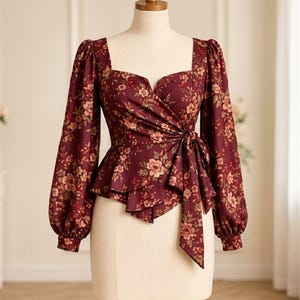 Könnte beinhalten: Eine langärmelige Wickelbluse aus tiefburgunderfarbenem Stoff mit Blumenmuster in Rosa- und Cremetönen. Die Bluse hat einen eckigen Ausschnitt, Puffärmel mit geknöpften Manschetten und einen seitlichen Bindegürtel an der Taille.