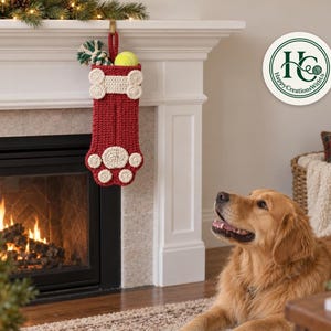 May include: A red crocheted pet stocking featuring a cream-colored bone design at the top and a paw print at the bottom. The stocking is filled with a rope toy and a yellow tennis ball, hanging from a white fireplace mantel.