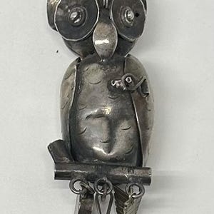 May include: A silver-toned owl pendant with detailed features. The owl is perched on a branch, with dangling charms below. The pendant has a textured surface and a vintage aesthetic, suitable for jewelry or craft projects.
