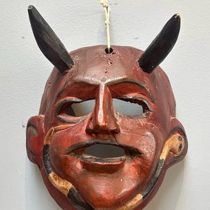 May include: A red wooden mask with black horns and a snake design around the mouth. The mask has a carved face with a wide mouth and large eyes.