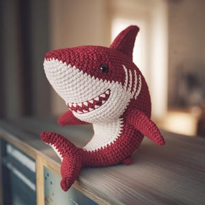 May include: A crochet shark plush toy featuring a deep red body with a contrasting white underbelly, snout, and fin tips. The shark has a wide, smiling mouth with visible white teeth, a single black eye, and a prominent dorsal fin.