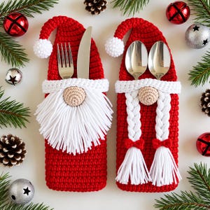 Könnte beinhalten: Zwei rote Bestecktaschen aus Häkelarbeit in Form von Weihnachtsmannmützen. Eine hat einen weißen Bart und enthält eine silberne Gabel und ein Messer. Die andere hat zwei weiße Zöpfe mit roten Schleifen und enthält zwei silberne Löffel.