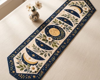 May include: A rectangular quilted table runner featuring a central moon phase sequence in gold and dark blue, surrounded by green foliage and white flowers. The border is dark blue with gold star accents, and the fabric has a stitched texture.