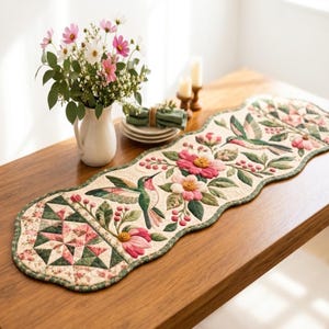 May include: A quilted table runner featuring a scalloped edge, decorated with embroidered hummingbirds, pink and white flowers, and green leaves. The runner includes a geometric star pattern at the end and a cream-coloured background.