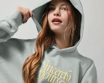 Może przedstawiać: Jasnozielona bluza z kapturem w kolorze szałwiowym z napisem Sincerely Yours wyhaftowanym jasnym żółtym kursywą na piersi. Odzież posiada kaptur z pasującymi sznurkami oraz ściągacze przy długich rękawach.
